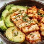 A vibrant bowl featuring crispy, golden-brown chicken pieces topped with a creamy, spicy Bang Bang sauce. Slices of colorful bell peppers and fresh greens surround the chicken, all arranged in a stylish white ceramic bowl on a rustic wooden table.