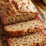 A moist, golden-brown banana bread loaf sitting on a rustic wooden surface, sliced to reveal a tender, moist interior dotted with ripe banana pieces, with a buttery crust and a few banana slices garnishing the top for contrast.