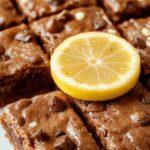 A vibrant display of lemon brownies arranged on a white plate, showcasing their bright yellow color with a slightly caramelized top and a fudgy, moist interior. The brownies are garnished with a dusting of powdered sugar and a few fresh lemon slices, contrasting beautifully with their golden crust and moist crumb.
