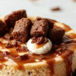 A decadent caramel brownie cheesecake topped with a drizzle of caramel sauce and a sprinkle of chopped nuts. The cheesecake has a rich, creamy texture with a dark chocolate crust visible around the edges. Garnished with a few dollops of whipped cream and delicate chocolate shavings, it is plated on a rustic wooden surface with a fork beside it, inviting indulgence.