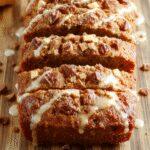 A slice of golden pumpkin bread with a crumbly streusel topping and a glossy maple glaze drizzled on top. The bread is placed on a rustic wooden board, with a soft focus on the textured streusel and moist interior showing through the cut edge. The colors are warm and inviting, featuring rich orange hues, golden brown streusel, and a hint of maple syrup shine.