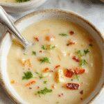 A steaming bowl of vibrant green and orange spring vegetable soup garnished with fresh herbs, served in a rustic white bowl on a wooden table, with colorful fresh vegetables surrounding it, showcasing a smooth, creamy texture and hints of herbs and spices.