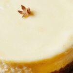 A slice of Lemon Cheesecake Cake on a white plate, topped with fresh lemon zest and a drizzle of lemon glaze. The cake has a creamy, golden-yellow filling with a smooth texture and a graham cracker crust. The background features whole lemons and a rustic wooden surface, highlighting the fresh citrus theme.