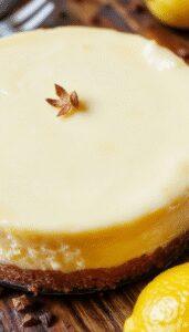 A slice of Lemon Cheesecake Cake on a white plate, topped with fresh lemon zest and a drizzle of lemon glaze. The cake has a creamy, golden-yellow filling with a smooth texture and a graham cracker crust. The background features whole lemons and a rustic wooden surface, highlighting the fresh citrus theme.