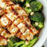 A vibrant bowl featuring grilled chicken slices nestled atop steamed broccoli florets, drizzled with a creamy garlic sauce. The dish is garnished with fresh herbs and served on a rustic wooden table, showcasing the colorful ingredients and inviting textures.