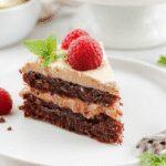 A slice of dark chocolate raspberry mousse cake on a white plate, showcasing layered textures with glossy dark chocolate glaze, ripe raspberries on top, and a light dusting of cocoa powder. The cake’s rich interior reveals a smooth, airy mousse with hints of berries, complemented by a delicate crumb crust, all beautifully presented against a neutral background.