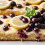A slice of vibrant blue blueberry sheet cake on a white plate, topped with fresh blueberries and a light dusting of powdered sugar, with a golden-brown crust and moist interior visible. The cake is garnished with sprigs of mint and set on a rustic wooden table, evoking a fresh, summery feel.