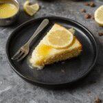 A slice of vibrant yellow lemon custard cake with a golden-brown crust on a rustic white plate, garnished with fresh lemon slices and mint leaves, with a background of a wooden table and soft natural lighting, emphasizing the cake’s moist texture and bright citrus flavor.