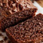 A slice of moist chocolate pumpkin bread on a rustic wooden plate, showing its rich dark chocolate swirls and bright orange pumpkin streaks, topped with a light dusting of powdered sugar. The bread’s crumb looks tender and dense, with a glossy, slightly shiny crust, and the background features a cozy kitchen setting with fall-themed decor.