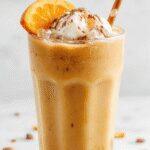 A tall glass filled with a creamy, light-colored pumpkin chai iced latte topped with a swirl of whipped cream and a sprinkle of cinnamon, garnished with a cinnamon stick. The beverage showcases a smooth, frothy texture with visible specks of spice, served on a rustic wooden table with autumnal decor in the background.