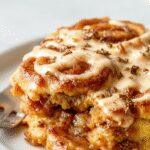A golden-brown pumpkin cinnamon roll bake sliced to reveal swirls of cinnamon and pumpkin filling, topped with a light glaze and sprinkled with cinnamon, plated on a rustic white dish with a cozy kitchen background.
