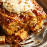 A golden-brown pumpkin French toast casserole topped with a dusting of powdered sugar and fresh maple syrup. The casserole is garnished with sliced bananas and a few sprigs of mint, served in a rustic ceramic dish. The background features a wooden table and a cozy breakfast setting with a glass of orange juice and a fork.