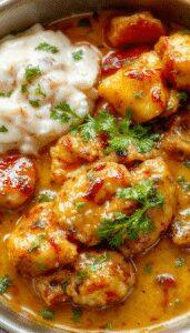 A creamy serving of homemade butter chicken garnished with fresh cilantro, placed on a rustic white plate. The rich orange sauce contrasts with the green herbs, with tender chicken pieces visible under the glossy coating. The background features a wooden table, and a side of steamed rice and naan bread add to the inviting presentation.