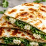 A close-up of a golden-brown quesadilla filled with vibrant green spinach and white crumbled feta cheese, sliced to reveal the gooey melted cheese and fresh spinach inside. The quesadilla is plated on a rustic wooden surface with a few fresh spinach leaves scattered around, highlighting the fresh ingredients and crispy texture.
