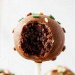 A close-up of a vibrant plate featuring multiple homemade Starbucks-style cake pops with smooth chocolate coating and colorful sprinkles atop a rustic wooden table. The cake pops are evenly sized, showcased with a glossy finish, and the background features a soft-focus coffee cup and a cozy ambiance.