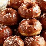 A close-up of golden-brown churro donut bites arranged on a rustic white plate. The bites are coated with cinnamon sugar, showcasing their crunchy exterior. In the background, a small bowl of apple compote and a sprinkle of cinnamon create a tempting scene. The vibrant colors of caramelized apple pieces add warmth and appeal, highlighting the crispy and fluffy textures of the bites.
