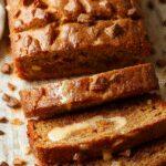 A sliced piece of pumpkin bread topped with a swirl of vanilla pudding and a sprinkle of cinnamon, set on a rustic wooden platter with a spoon nearby. The bread's golden crust contrasts with the creamy vanilla filling inside, and cinnamon dusting adds warmth to the presentation.