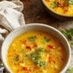 A vibrant bowl of vegan lemon lentil soup garnished with fresh herbs, bright yellow in color, served on a rustic wooden table with lemon wedges and a sprinkle of turmeric on top. The smooth, hearty soup showcases lentils and a zesty lemon zest twist, inviting a warm and wholesome feeling.