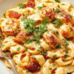 A close-up of a creamy pasta dish topped with crispy bacon and tender chicken pieces, garnished with chopped herbs. The pasta is served on a white plate with a fork beside it, highlighting the rich texture and vibrant colors of the ingredients.