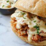 A close-up of a hearty Chicken Alfredo Sloppy Joe sandwich on a toasted hoagie roll, topped with melted cheese and creamy Alfredo sauce, garnished with chopped parsley. The sandwich is partially sliced, revealing tender ground chicken and rich white sauce inside, set on a rustic wooden plate.