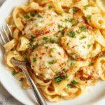 A creamy chicken pasta dish featuring tender slices of chicken breast coated in a rich garlic Parmesan sauce, served on a white plate with a sprinkle of chopped parsley and grated Parmesan cheese. The pasta is coated in a smooth, buttery sauce with visible garlic bits and a golden hue, alongside a side of fresh green herbs and a lemon wedge for garnish, creating an inviting and appetizing presentation.