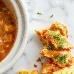 A creamy buffalo chicken dip served in a white ceramic bowl, topped with chopped green onions and sprinkled with shredded cheddar cheese. The dip is surrounded by celery sticks and carrot slices on a rustic wooden table, with a spoon resting beside the bowl. The vibrant orange buffalo sauce and melted cheese create an appetizing contrast, highlighting its creamy texture and inviting presentation.