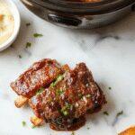 A plate of succulent beef short ribs glazed with a rich, glossy sauce, garnished with fresh herbs. The ribs are tender, falling apart, with visible boneless meat and a caramelized exterior. They are served alongside steaming noodles and colorful roasted vegetables, presented on a rustic wooden table with a cozy, inviting atmosphere.