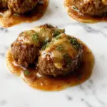 A close-up shot of golden-brown French onion meatballs arranged on a rustic white plate, topped with melted cheese and garnished with caramelized onion slices. The savory onion sauce glistens in the background, creating a rich and inviting scene.