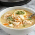 A steaming bowl of Greek Lemon Chicken Soup featuring tender shredded chicken, vibrant lemon slices, fresh herbs, and a broth with a bright yellow hue. The soup is garnished with fresh parsley and lemon wedges, served in a rustic white bowl on a wooden table, with a spoon resting beside it. In the background, there are additional ingredients like lemons, herbs, and chopped vegetables, creating a warm and inviting atmosphere.