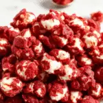 A close-up of vibrant red velvet popcorn drenched in a glossy red syrup, piled high in a spooky black bowl. The popcorn appears glossy, with some pieces coated more heavily, creating a hauntingly vivid red color. The background is dark and moody, with hints of eerie lighting emphasizing the sinister, blood-like appearance of the snack, perfect for a Halloween party.