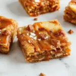 A close-up of salted caramel apple bars showcasing a golden-brown crust topped with glossy caramel, sliced fresh apples, and a sprinkle of sea salt. The bars are neatly cut and arranged on a rustic wooden surface, with caramel drizzles and apple slices visible, highlighting their layered texture and inviting appearance.