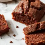 A freshly sliced loaf of moist chocolate pumpkin bread reveals a rich, dark interior speckled with chocolate chips. The bread is beautifully presented on a rustic wooden board, with a few slices fanned out to showcase the tender crumb and glossy chocolate streaks. The top is slightly cracked, hinting at its moist texture, with a dusting of cocoa powder around the edges. In the background, a cozy kitchen setting creates an inviting atmosphere, emphasizing the indulgent yet homemade feel of the baked good.