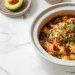 A hearty slow cooker taco casserole served on a rustic plate, featuring a vibrant mixture of seasoned ground beef, melted cheese, fresh tomato, and chopped green onions. The casserole is garnished with cilantro and sour cream, with a side of crispy tortilla chips and a lime wedge. The dish showcases a colorful, layered presentation with a golden-brown cheese crust on top, inviting and appetizing.