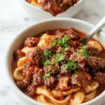 A rich, deep-brown beef ragu served over wide pasta noodles in a rustic white bowl, garnished with freshly chopped herbs, with steam gently rising, set on a wooden table with a rustic backdrop.