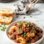 A rustic white bowl filled with rich beef ragu, showcasing tender shredded beef immersed in a thick, glossy tomato-based sauce with herbs. The dish is garnished with fresh basil leaves and served alongside al dente pasta on a wooden table, with steam gently rising to evoke warmth and comfort.