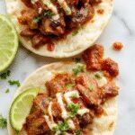 A serving of golden-brown birria tacos filled with tender, shredded beef topped with fresh cilantro and diced onions, drizzled with rich red consommé, presented on a rustic wooden plate with lime wedges and a side of chopped cilantro.