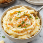 A close-up of a creamy, fluffy mound of mashed potatoes in a rustic white bowl. The potatoes are smooth with a subtle sheen, topped with a pat of melting butter and sprinkled with chopped chives. The background features a warm wooden table with festive fall decorations and a spoon resting beside the bowl, emphasizing comfort and holiday spirit.